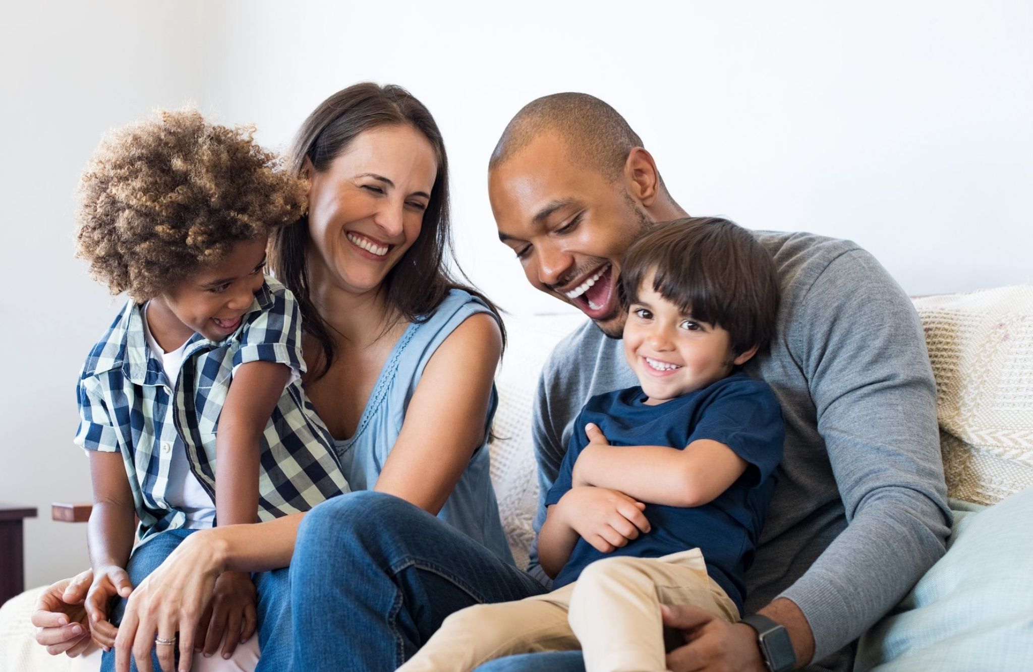 Hawthorne Northwest happy multiethnic family sitting on sofa laughing together. Cheerful parents playing with their sons at home. Black father tickles his little boy while the mother and the brother smile.